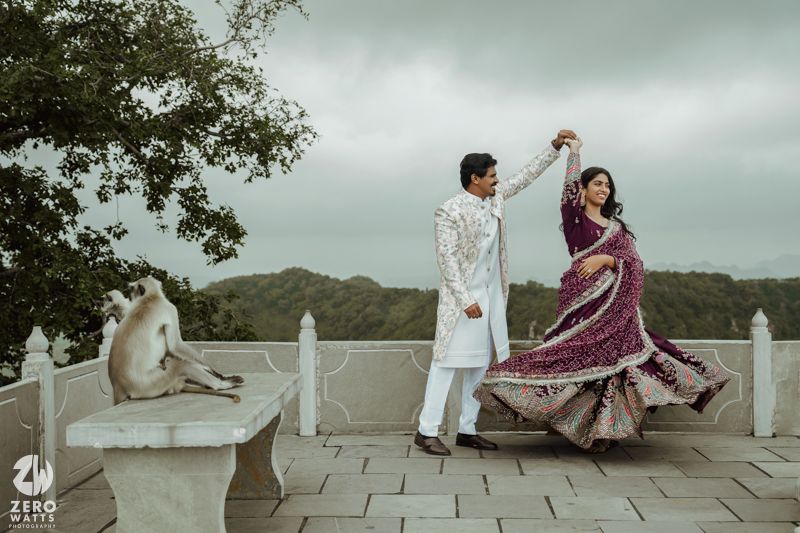 A couple in traditional attire creates romantic poses along a stone wall with lush greenery in the background.