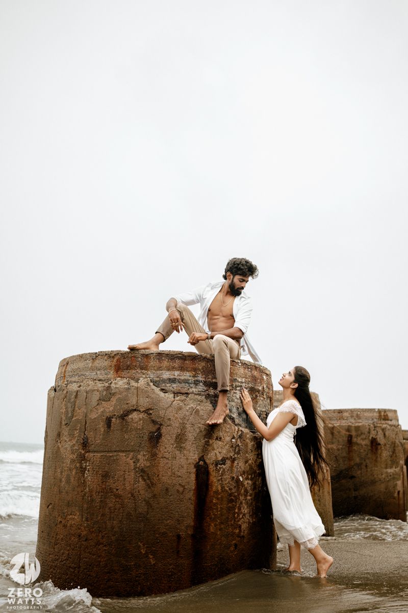 Man sitting atop a weathered stone pillar at beach while woman in white dress looks up at him adoringly.