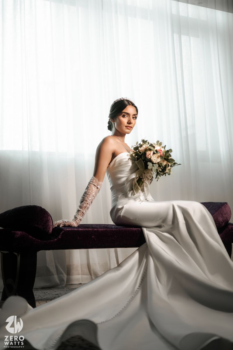 Bride in strapless white gown with lace gloves sits on velvet bench holding a floral bouquet.
