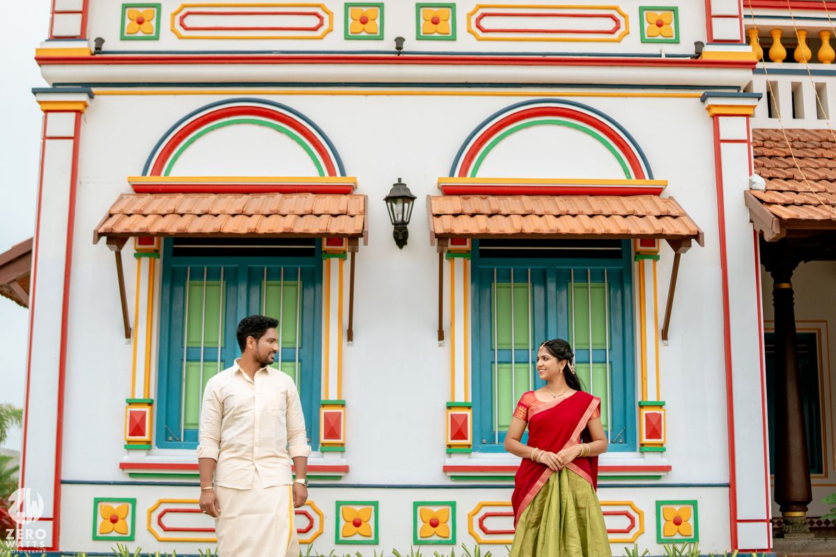 Couple in traditional South Indian outfits standing before colorful temple-style building with vibrant arched windows.
