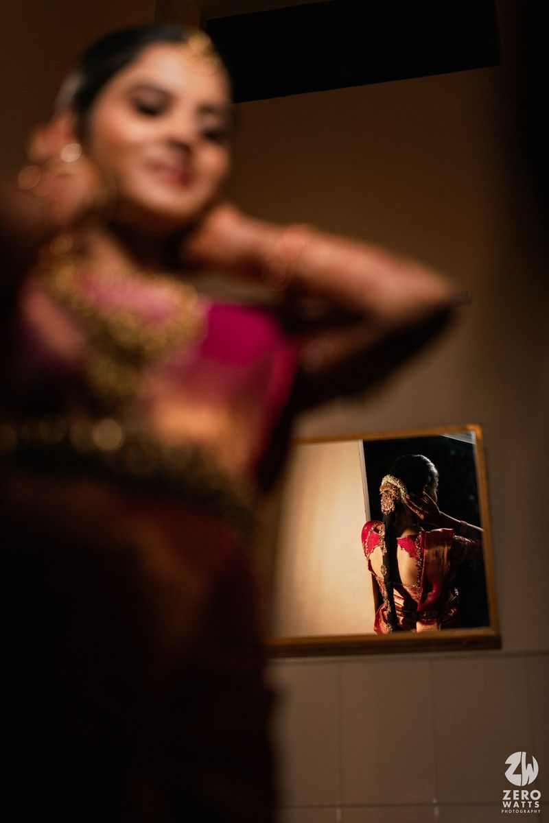 A bride in a pink saree adjusts her hair, with her reflection visible in a mirror during bridal preparations.