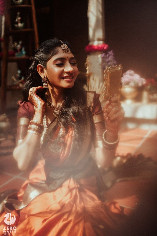 South Indian bride in orange silk saree with gold jewelry and maang tikka smiling during wedding ceremony.