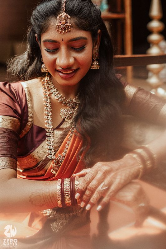 A smiling bride in traditional Indian bridal attire with jewelry and red saree looking down at her hands during wedding ceremony.