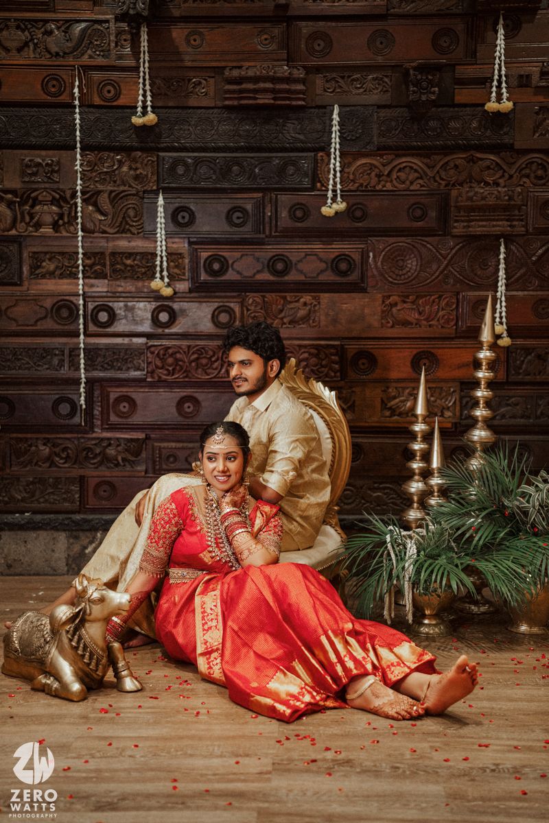 Indian couple in traditional wedding attire pose together on floor with carved wooden backdrop and brass lamps.