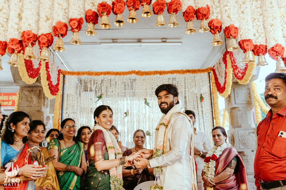 South Indian wedding ceremony with couple exchanging garlands under floral mandap with red and white decorations.
