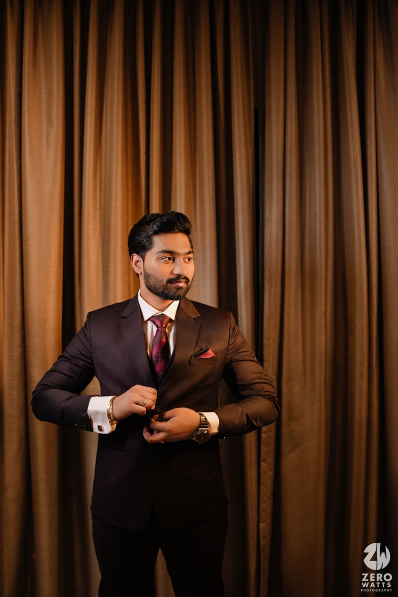 Formal portrait of groom in brown suit adjusting cufflink against bronze curtain backdrop.