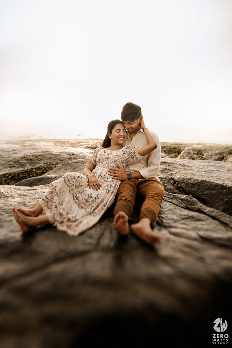 Couple lounging intimately on dark rocky terrain during a seaside photoshoot.