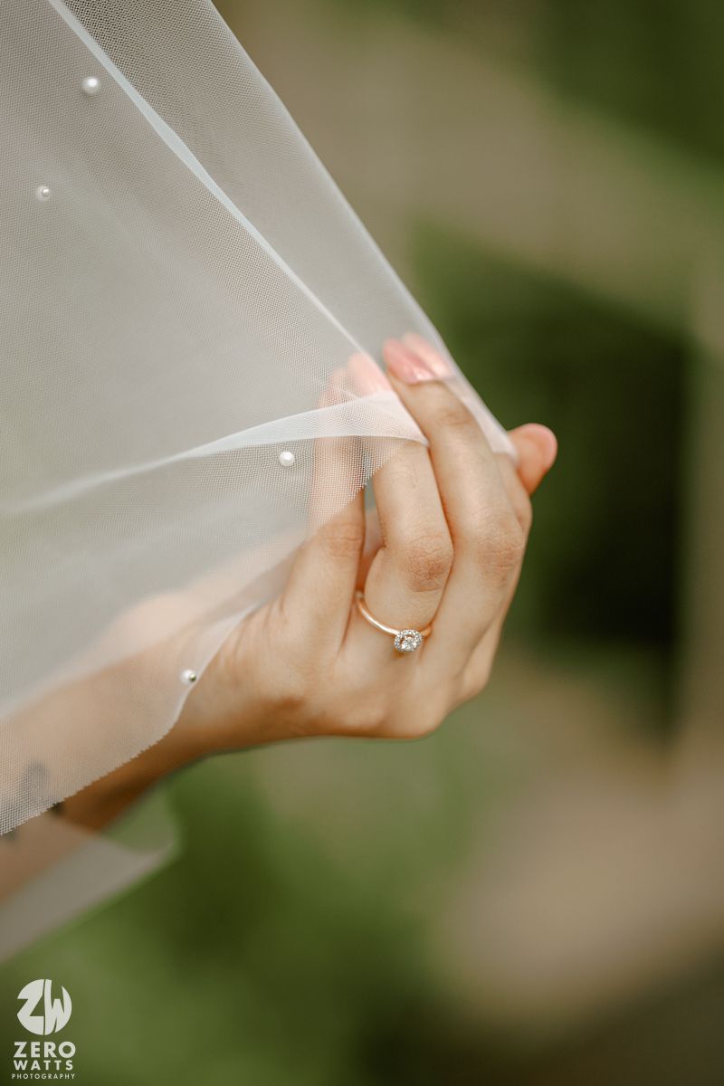 Close-up of hands holding delicate white wedding veil with engagement ring visible against blurred green background.