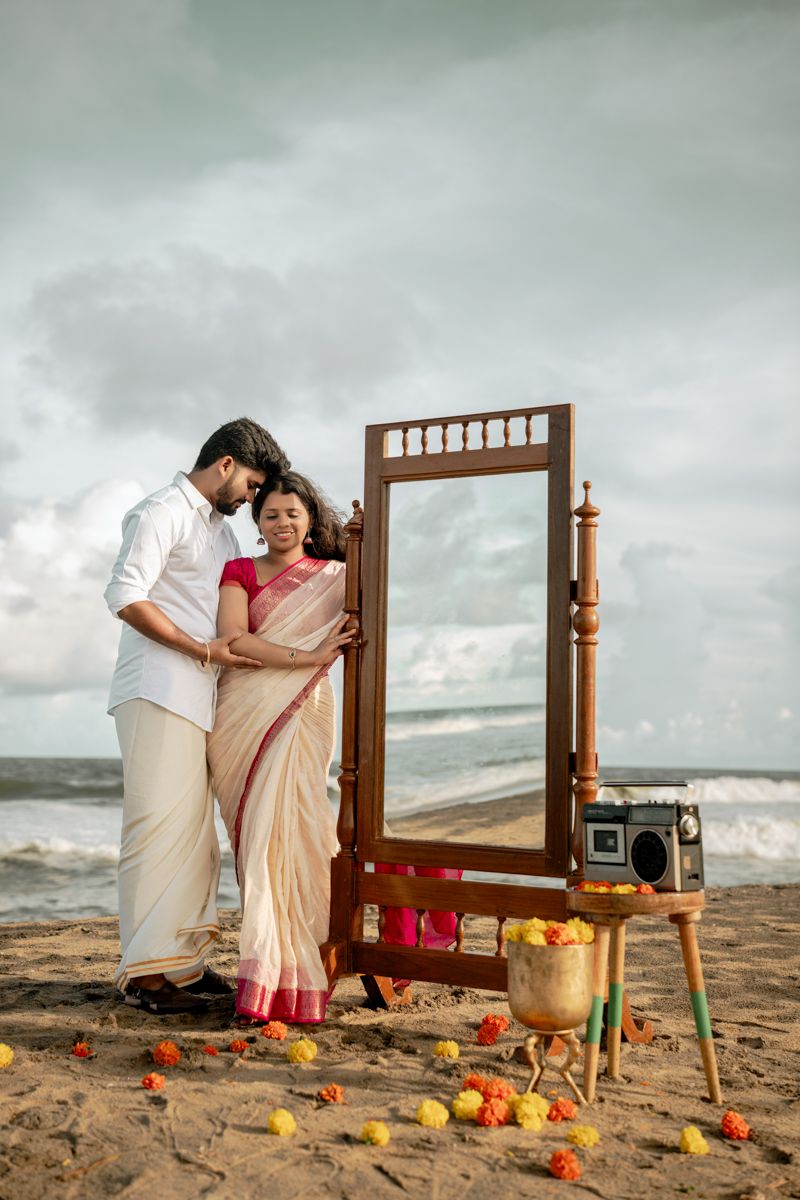 A romantic beach photoshoot setup with a vintage wooden mirror frame and colorful decorations on the sandy shore.