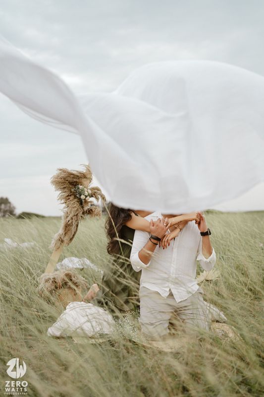 A couple in white clothing playfully embraces while lying in tall grass on a breezy summer day.
