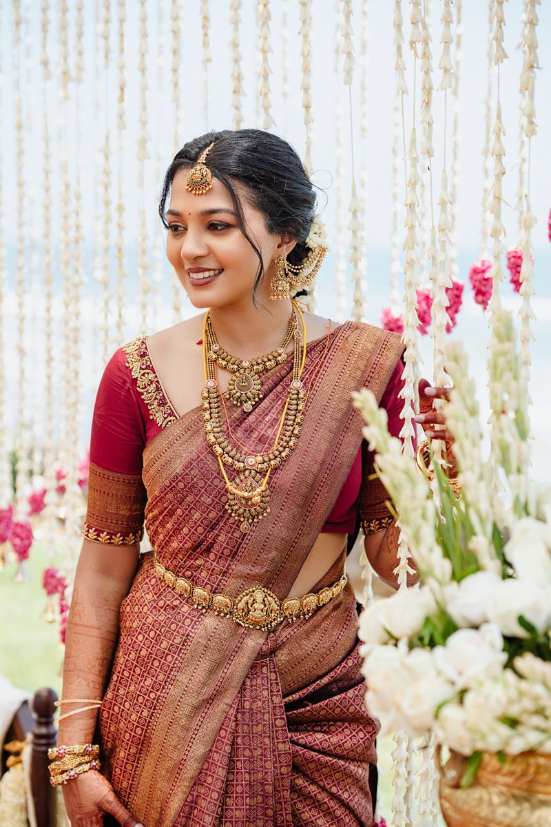 Elegant bridal attire featuring a maroon silk saree and ornate gold jewelry in a decorated setting.