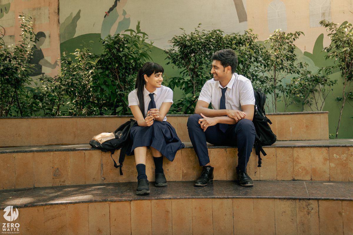 Two students in uniforms sit and chat on concrete steps surrounded by greenery.