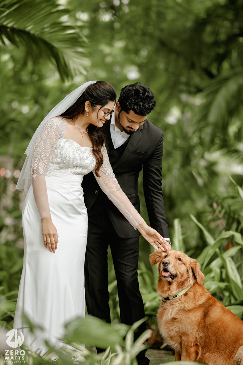 A couple in wedding attire pets a golden retriever dog in a lush green garden setting.