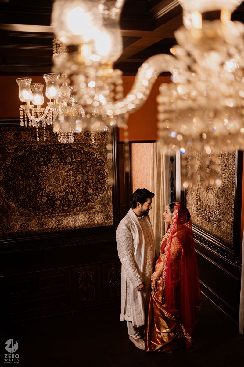 A couple in traditional Indian wedding attire stands beneath crystal chandeliers in an ornate room.