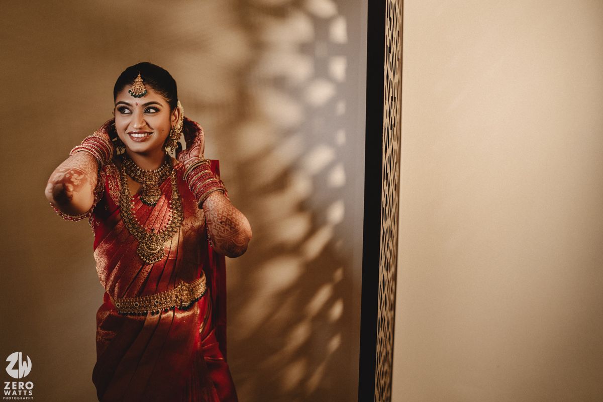 A bride in a traditional red and gold Indian wedding sari smiles while adjusting her earrings in soft lighting.