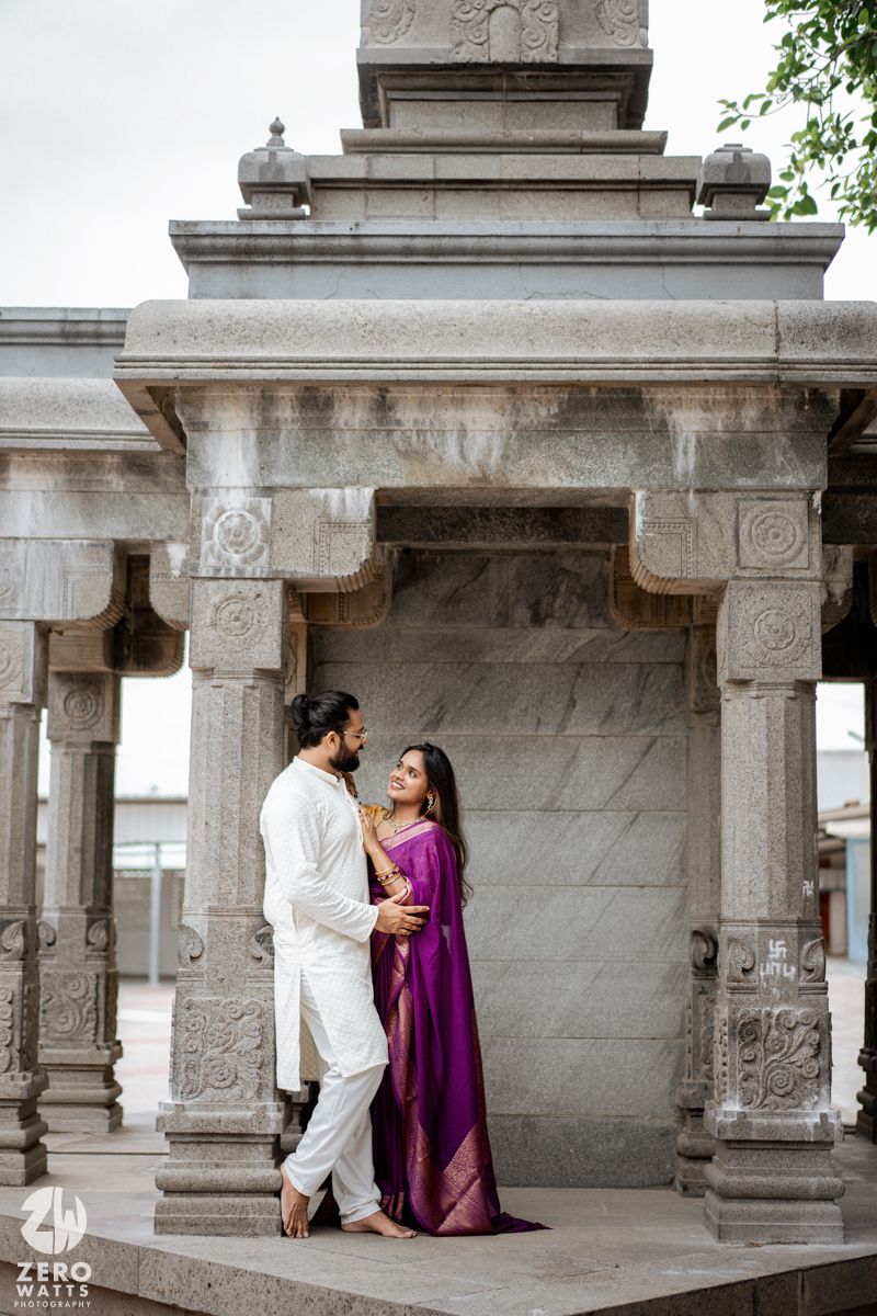Two people share an intimate moment in traditional Indian attire against stone temple architecture.