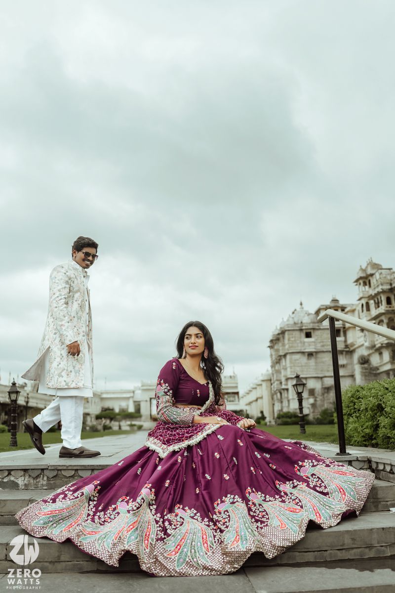 A couple in traditional Indian wedding attire poses dramatically on steps against a cloudy sky and palace backdrop.