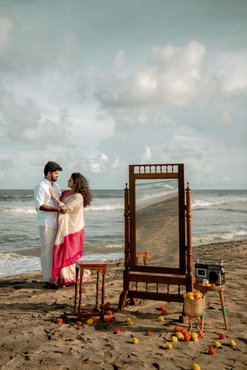 A couple shares a romantic moment on a beach with a wooden headboard and scattered pumpkins at sunset.