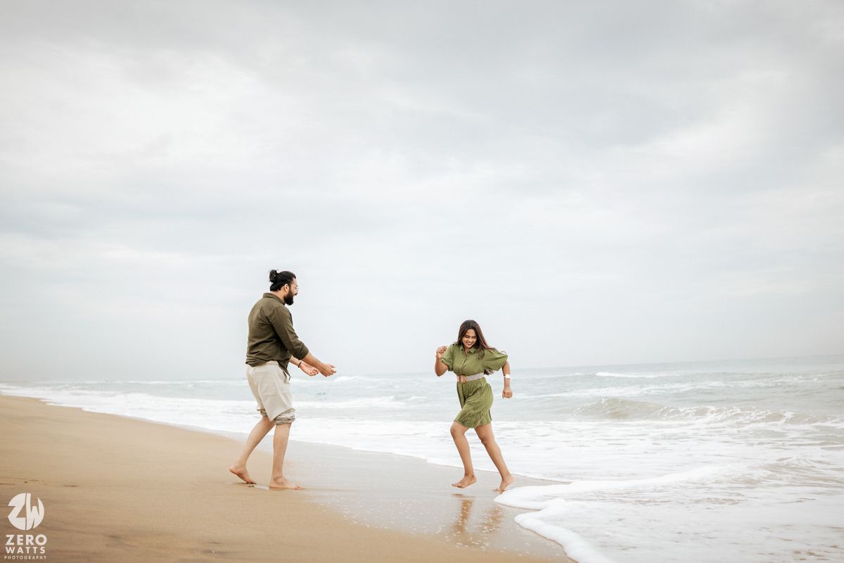 Two people playfully running and dancing together on a misty beach with waves crashing in the background.