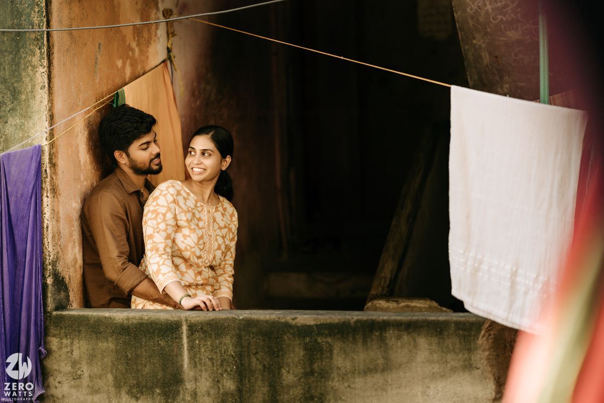 A couple shares a tender moment on a rustic balcony with hanging laundry and textured walls in warm lighting.