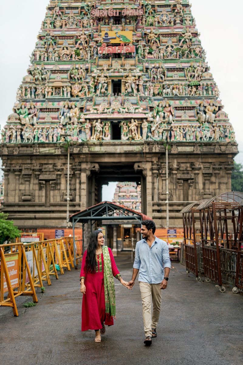 A couple walks hand in hand in front of an ornate Hindu temple with intricate carved architecture and sculptures.
