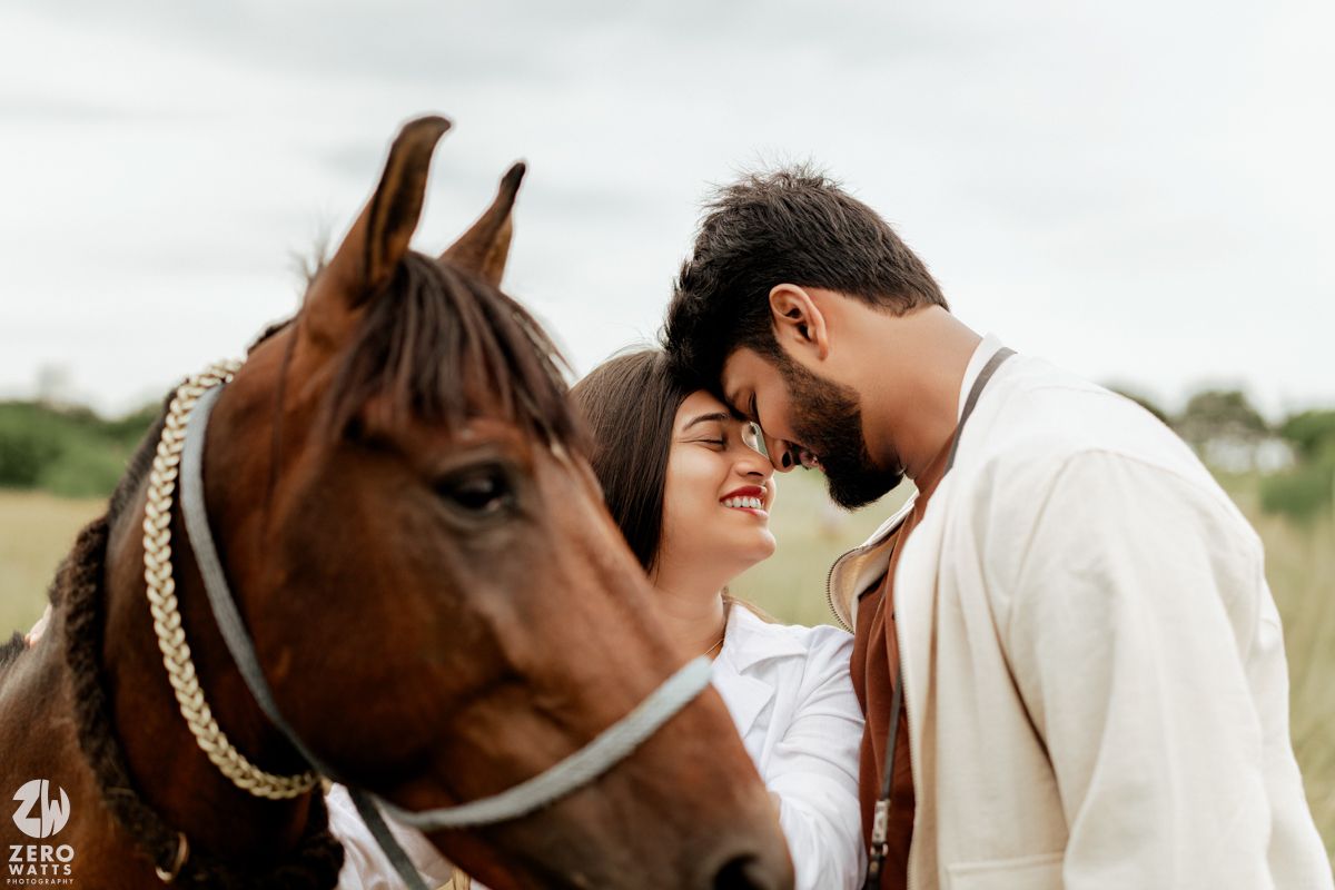 Couple interacts lovingly with a brown horse wearing a decorative bridle in a rustic field setting.