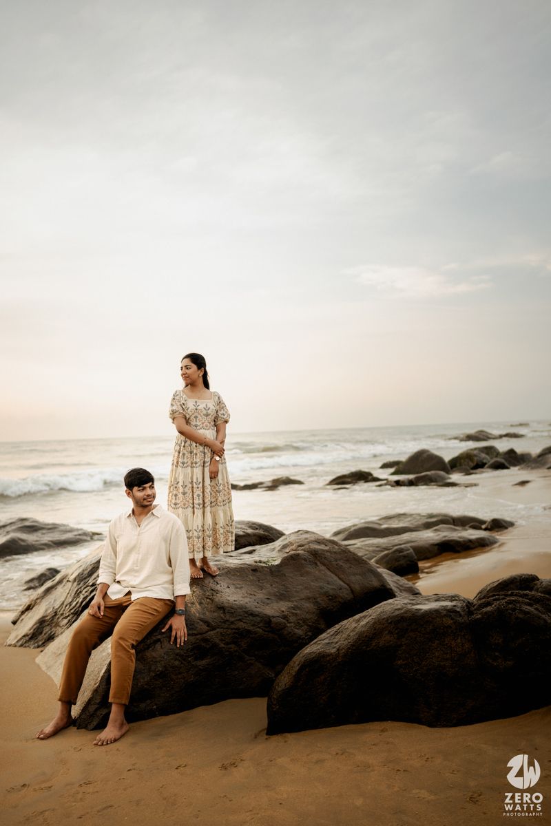 Couple sits peacefully on rocky coastal beach during sunset as waves crash against dark stones in the distance.