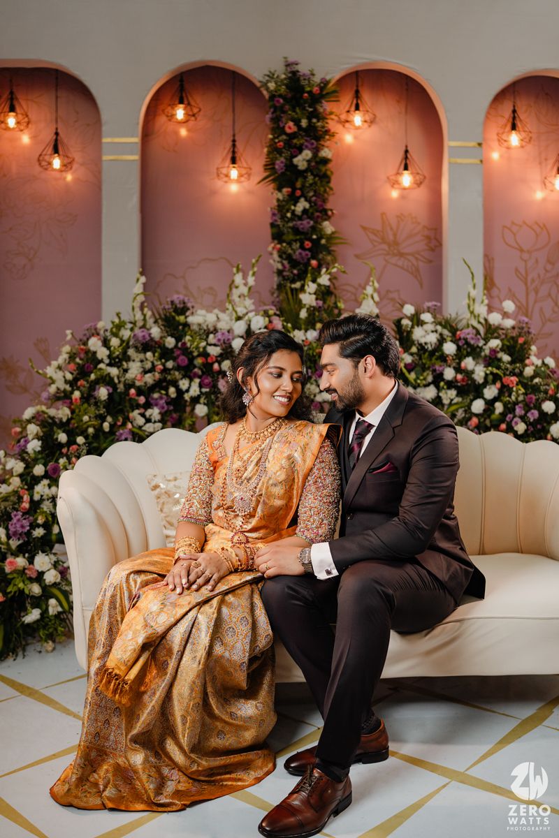 A couple sits together on a white sofa beneath arched walls adorned with floral arrangements and pendant lights.