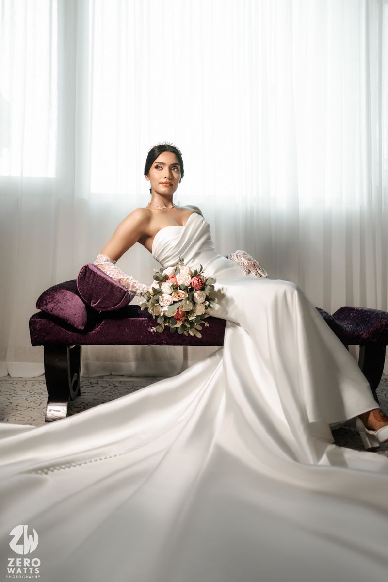 Bride in strapless white gown with lace gloves sits on velvet bench holding a floral bouquet.