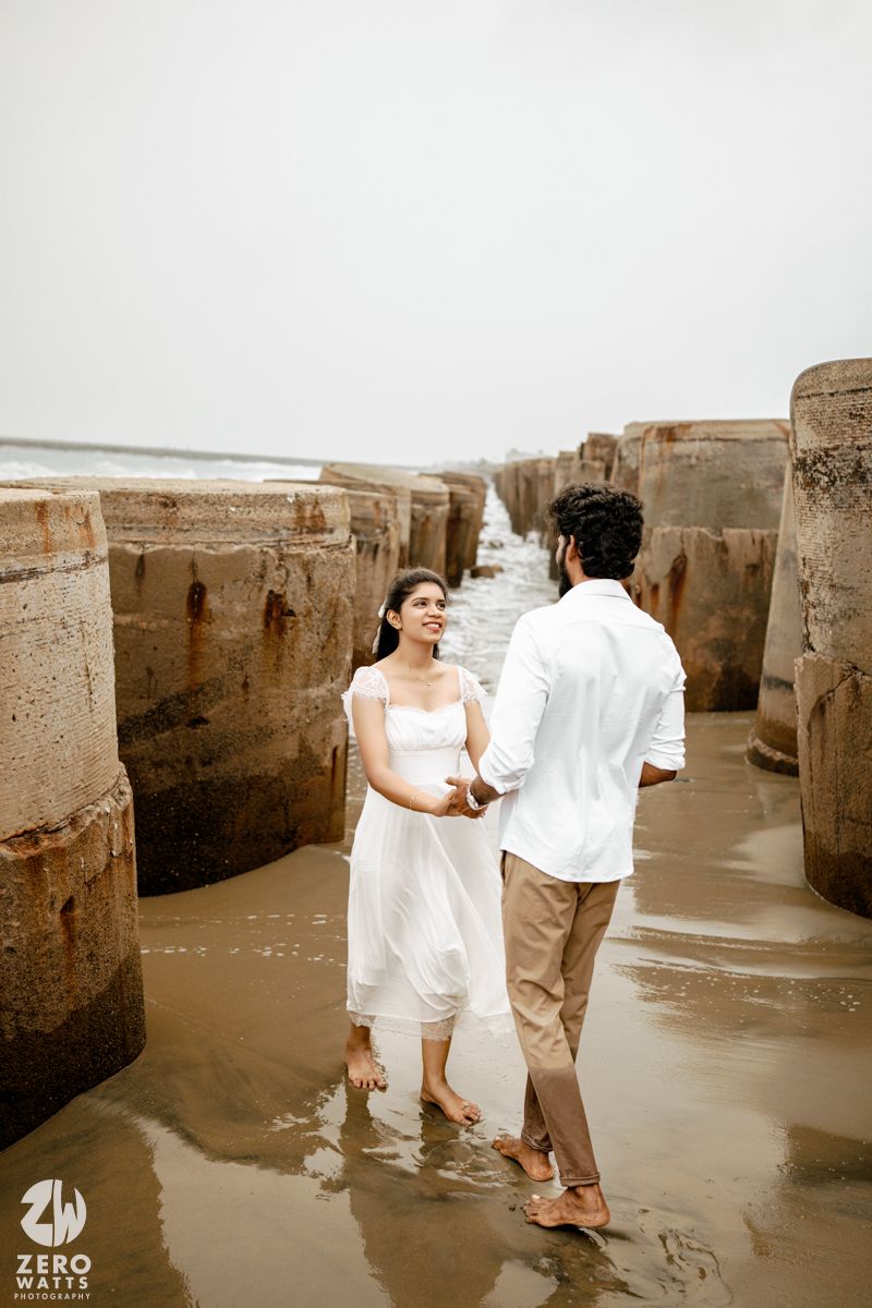 Couple in white outfits holding hands between concrete sea barriers on a wet beach, romantic pre-wedding photoshoot.