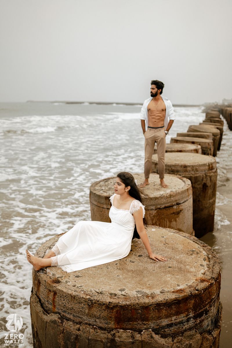 Woman in white dress poses on stone pier pillar while man in open shirt stands behind overlooking ocean waves.