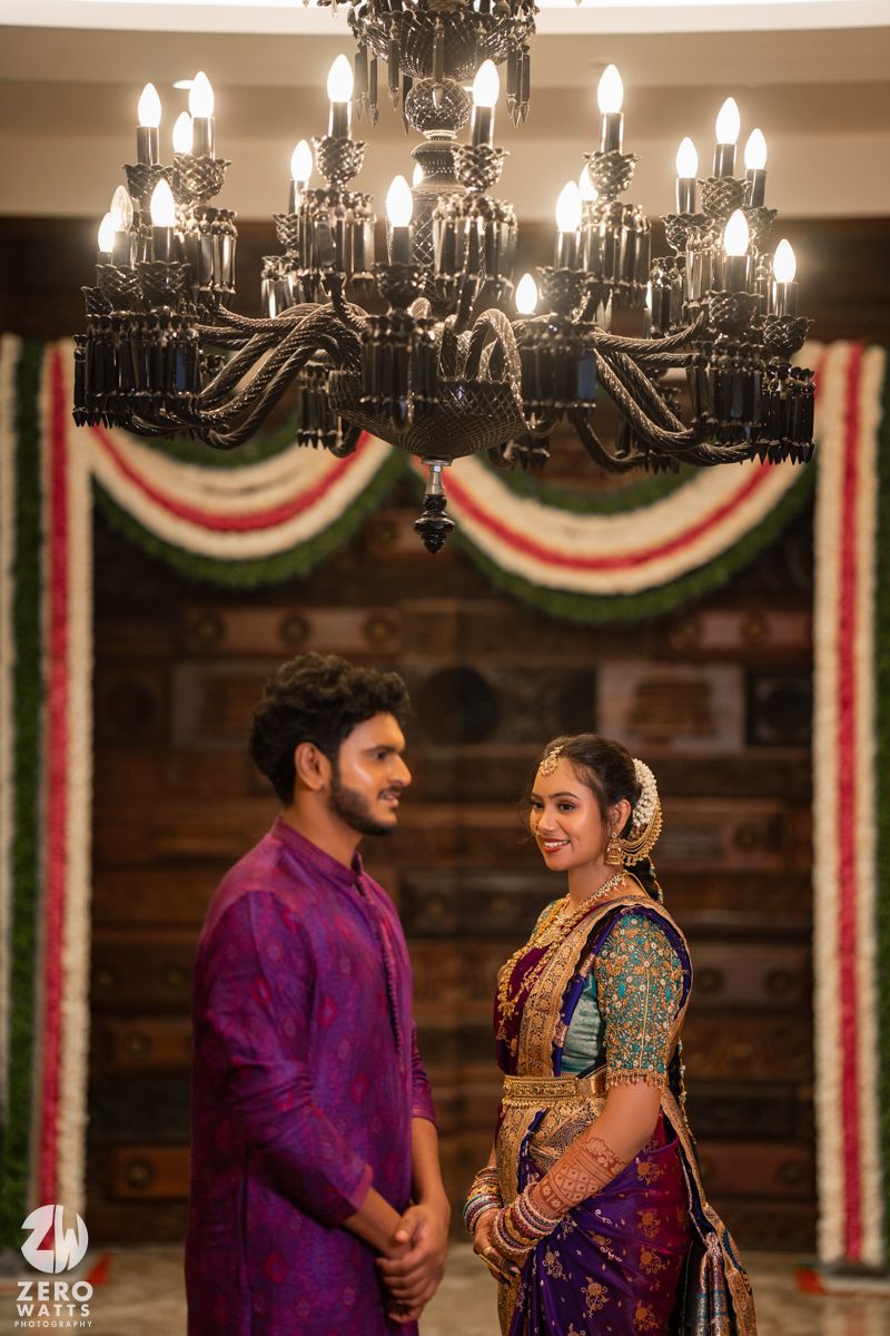 South Indian bride and groom in traditional attire standing under ornate chandelier with floral decorations.