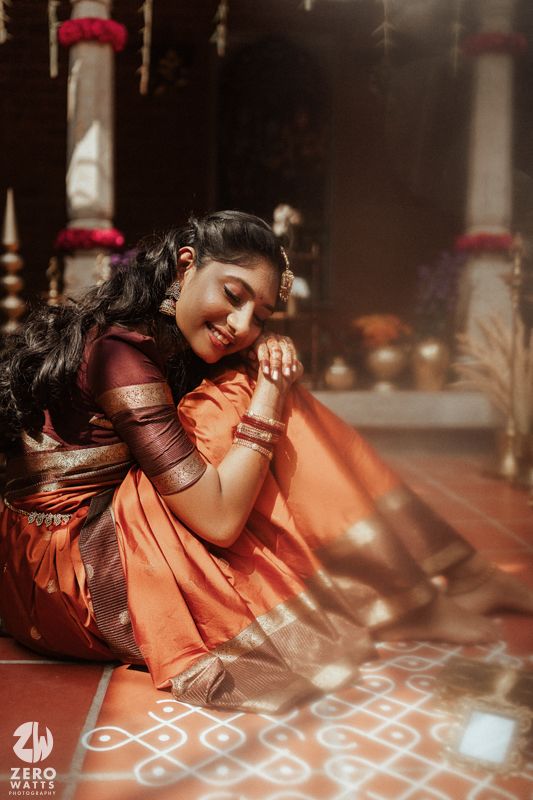 A South Indian bride in an orange Kanjivaram silk saree sits gracefully beside a kolam in soft sunlight.