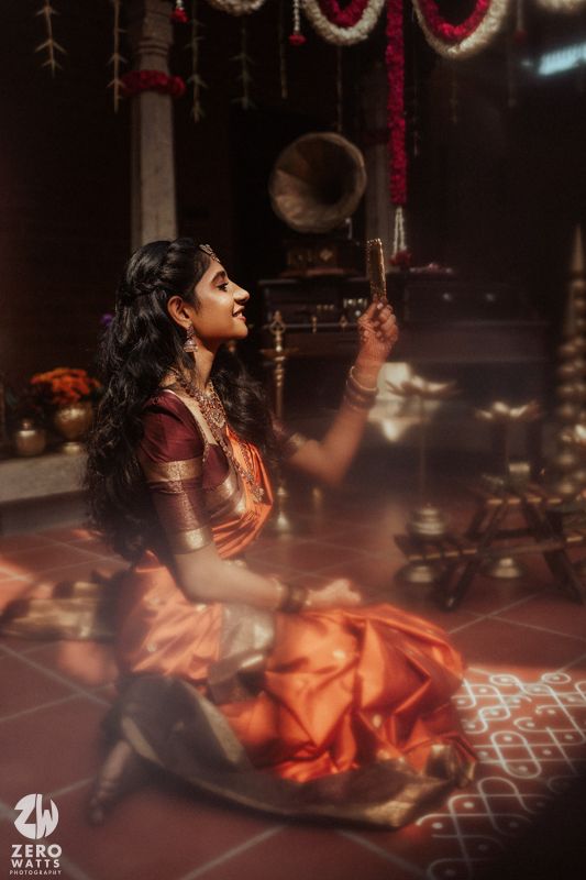 A woman in a vibrant orange and maroon silk saree sits gracefully on the floor, holding a diya in a traditional Indian setting.