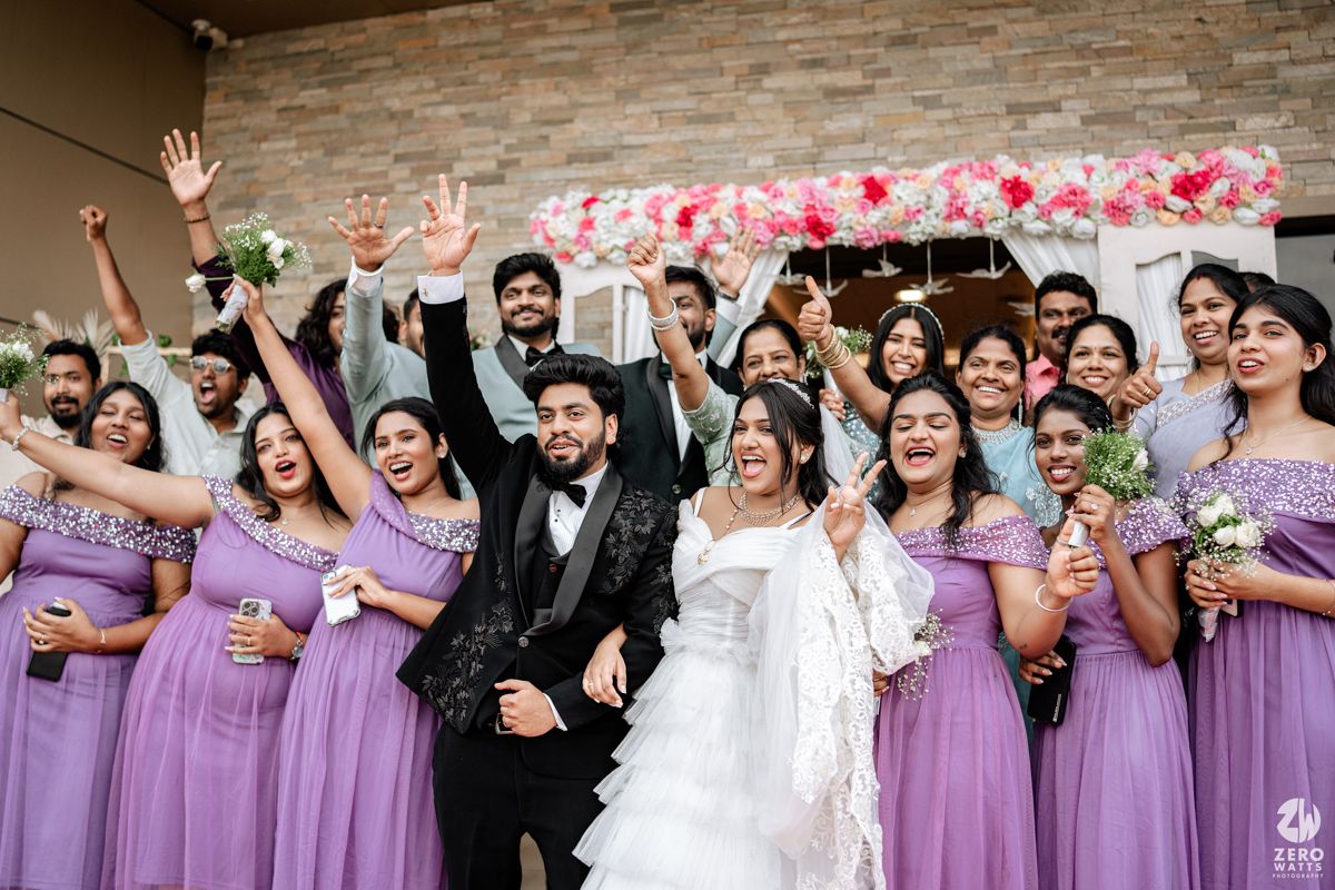 Joyful wedding party celebrates with bride in white and bridesmaids in lavender gowns raising hands outdoors.