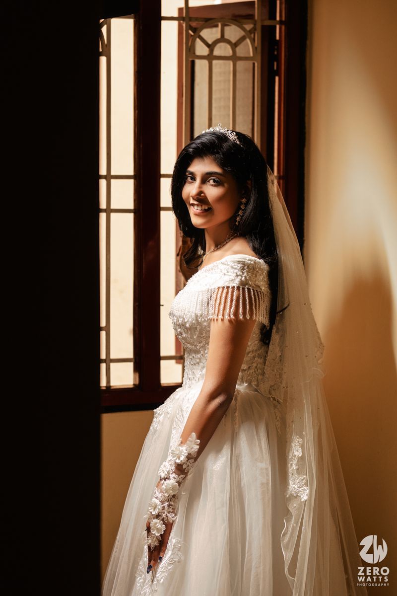 Bride in white off-shoulder lace wedding gown with veil poses by sunlit window, smiling elegantly.
