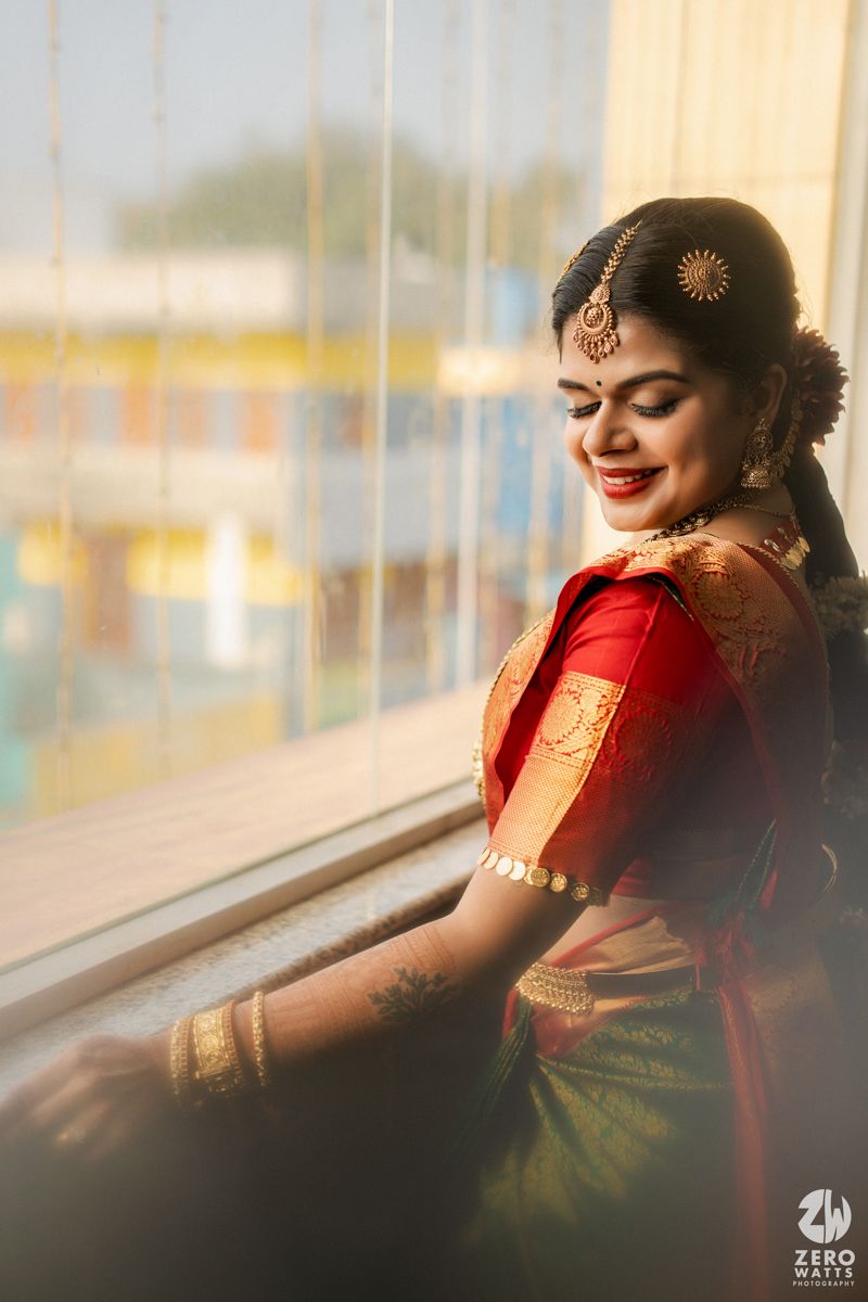 Smiling South Indian bride in red and green Kanjivaram saree with gold jewelry poses by sunlit window.