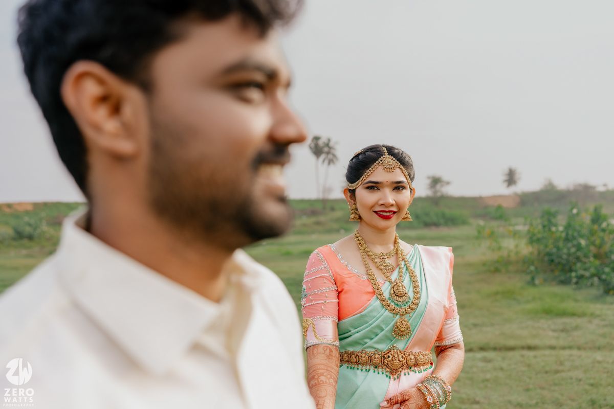 Bride in mint green saree with gold jewelry smiles as groom stands in foreground.
