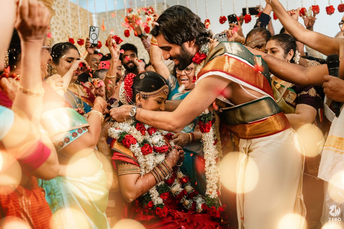 Traditional South Indian wedding ceremony with groom in dhoti exchanging garlands with bride in silk saree surrounded by guests.