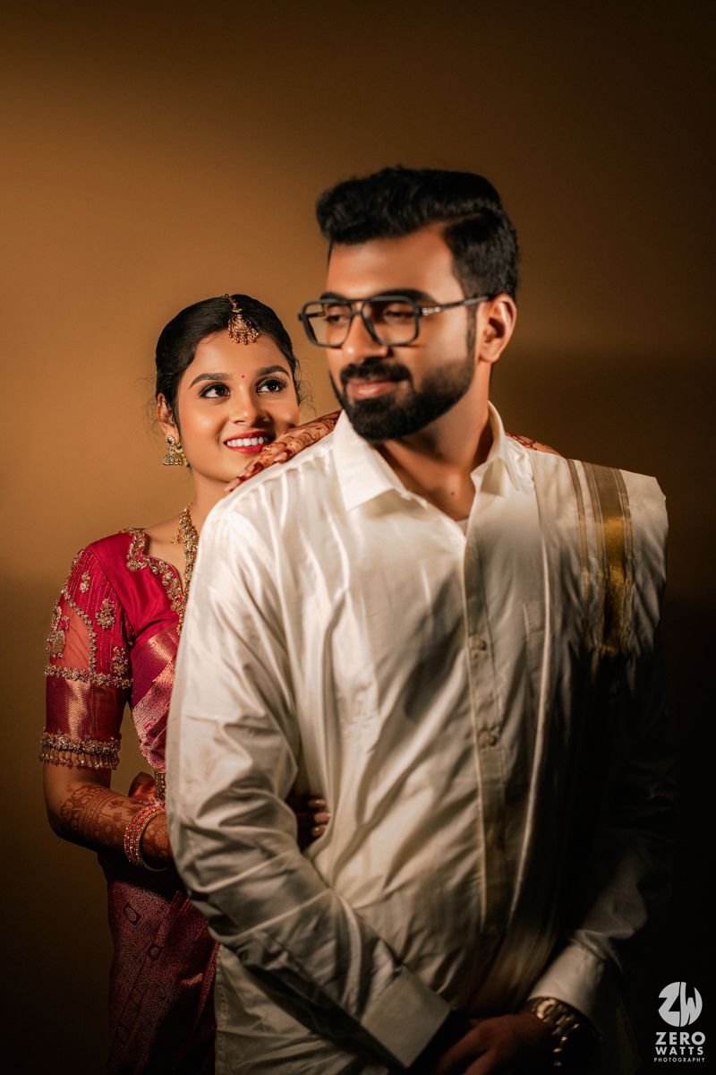 Indian bride in pink silk saree smiles behind groom in white traditional attire during South Indian wedding portrait.