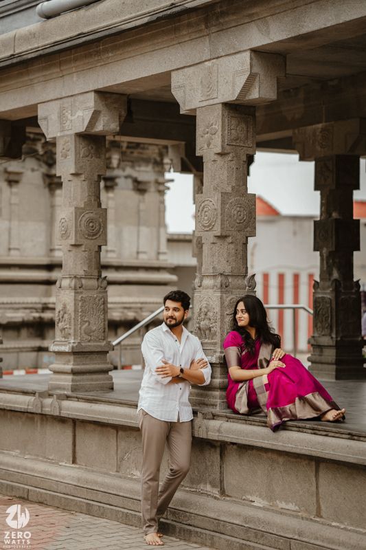 A couple in casual attire poses playfully among stone pillars and archways of a historic building.
