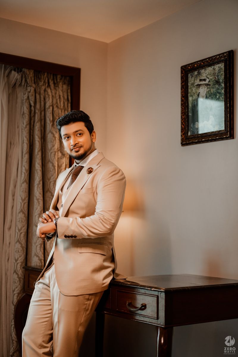 Elegant figure in beige suit stands confidently in room with framed artwork and curtains.