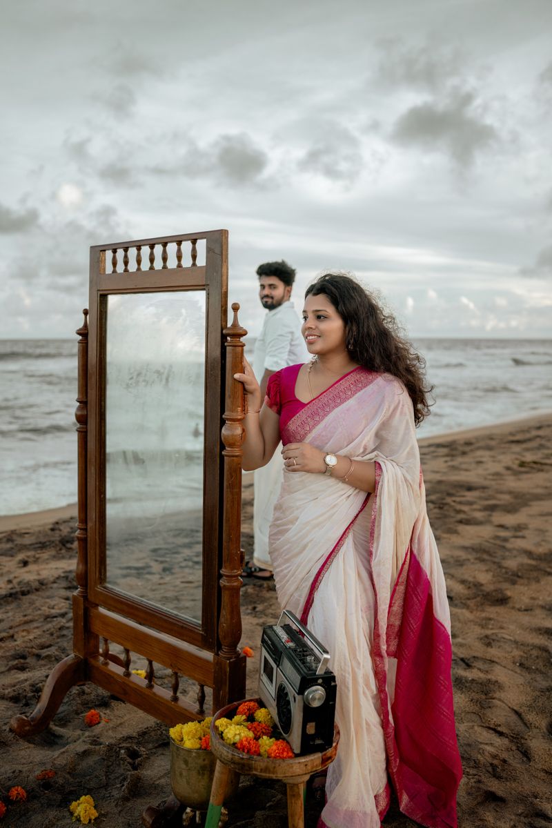 A person in a cream and pink saree poses artistically with a vintage mirror on a moody beach at sunset.