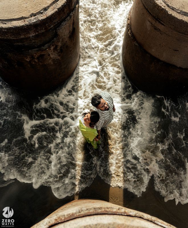 Waves crash dramatically around stone pillars at the beach with figures visible above.