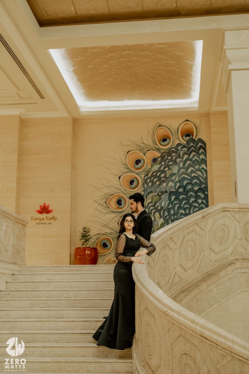 A couple in black outfits poses on an ornate white staircase with a decorative wall mural featuring peacock feathers.