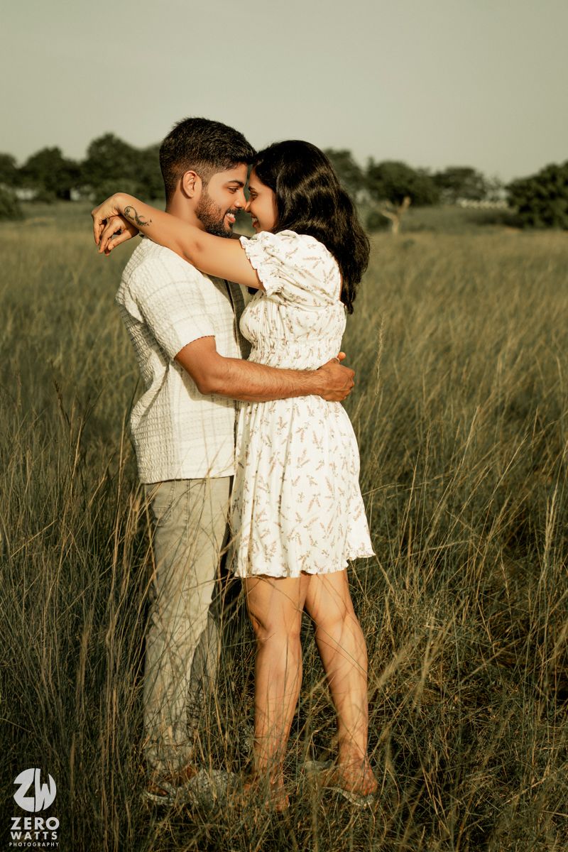 A romantic couple embraces in a grassy field wearing matching white outfits during a golden hour photoshoot.
