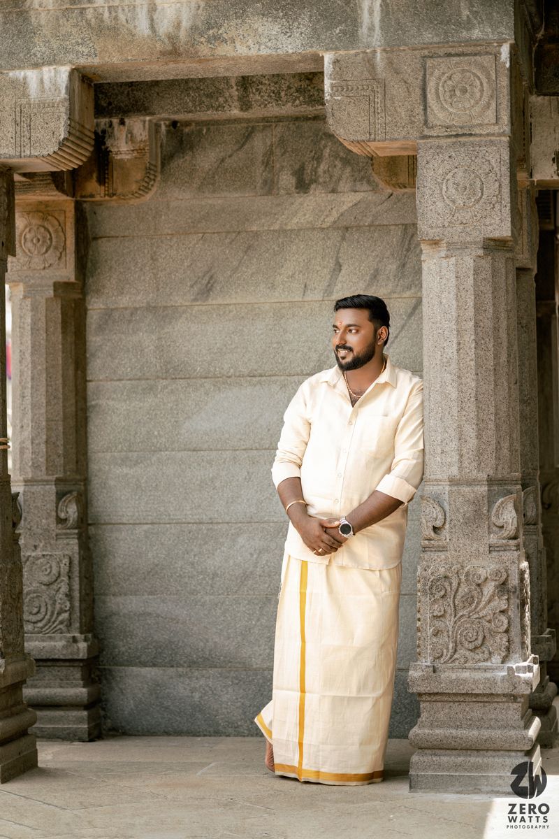 A person in traditional white and gold South Indian dhoti stands in an ancient stone temple corridor with carved pillars.