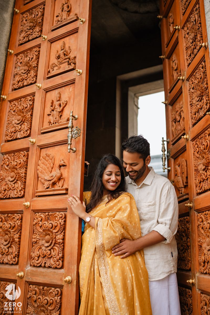 A couple shares intimate moments against ornately carved wooden doors in traditional Indian attire.