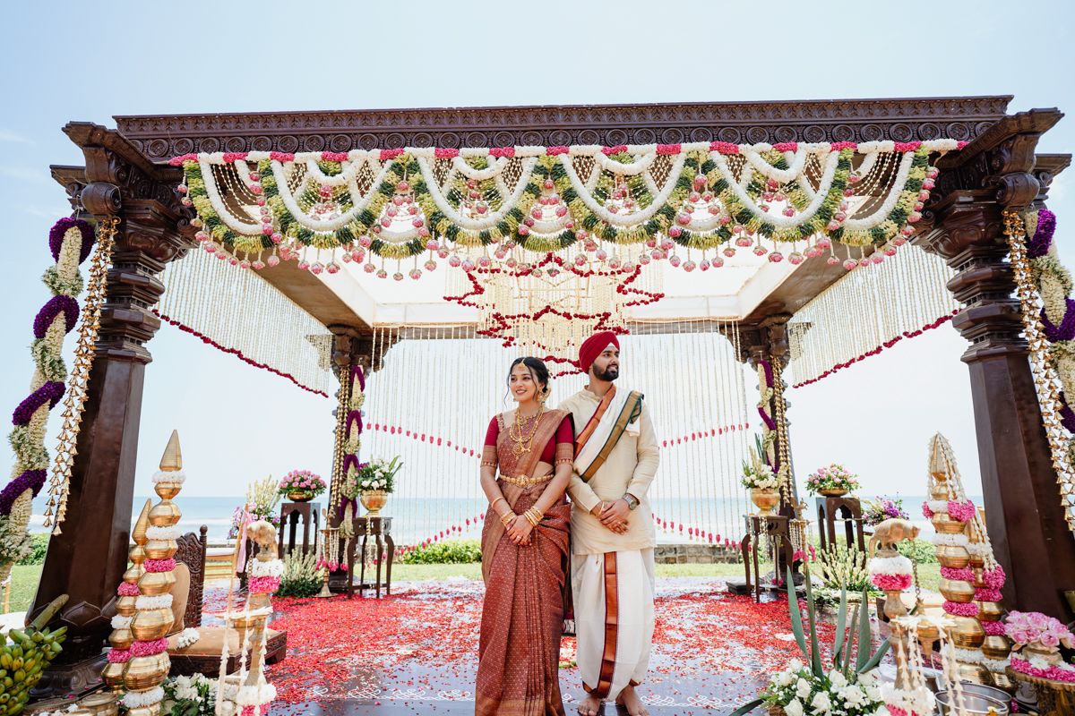Outdoor Indian wedding ceremony under ornate floral mandap with ocean backdrop and rose petals.