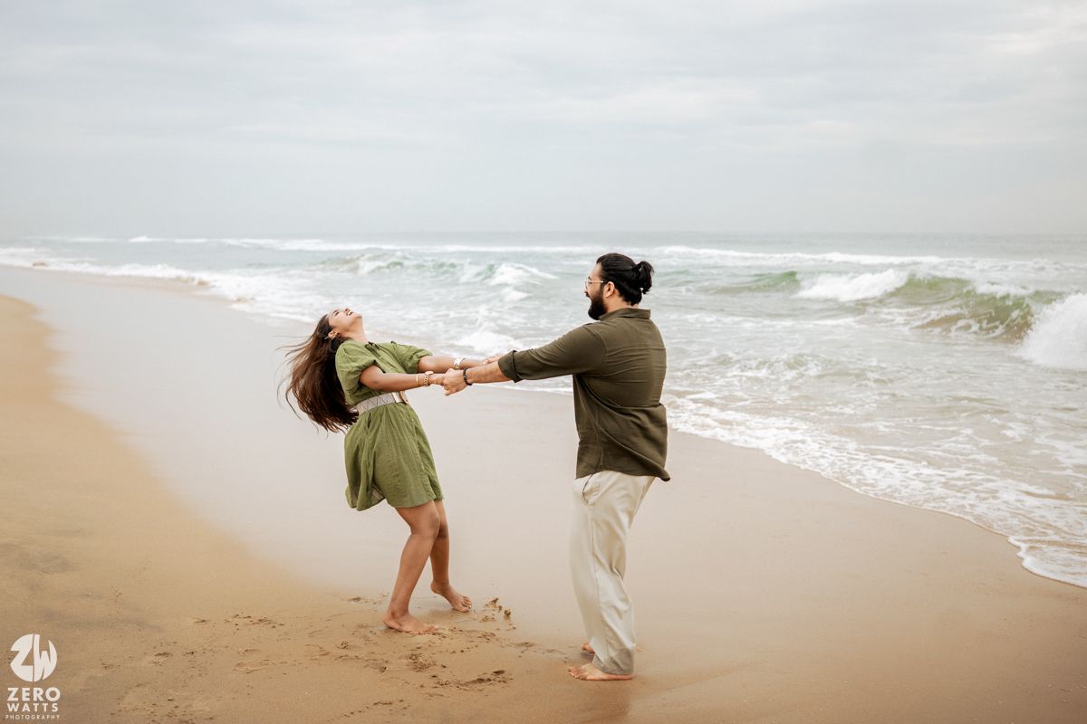 A couple in casual attire playfully walks along a misty beach shoreline with waves crashing behind them.